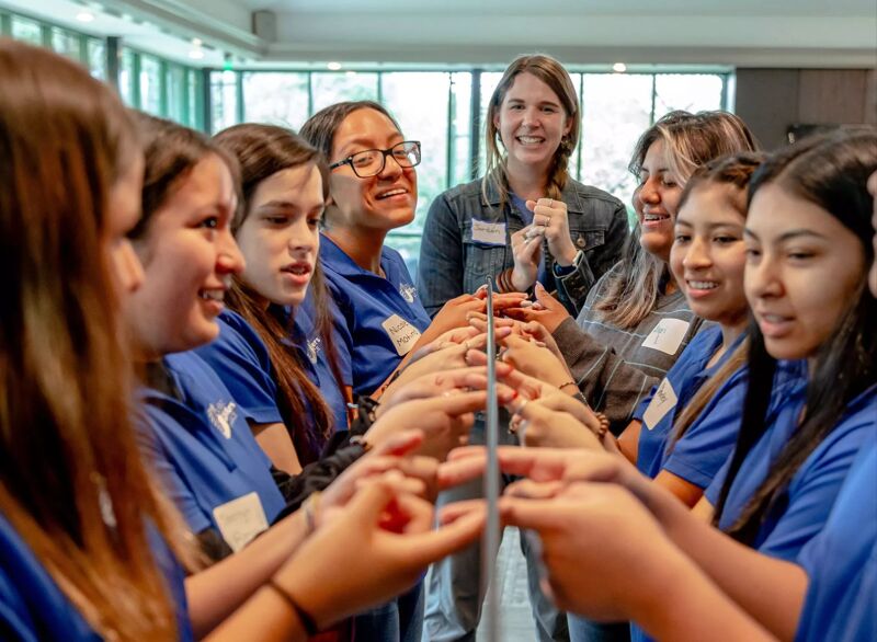 A group of young women, mostly wearing blue shirts, are gathered in a circle, extending their hands towards the center. An adult woman stands behind them, smiling and observing the activity. The setting appears to be indoors, possibly a classroom or a conference room, with natural light coming through the windows.